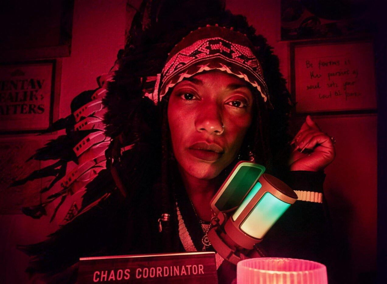Chief I'ni MichElle.magic in the red-lit Shadow Lounge studio, wearing a feathered headdress beside the microphone and a Chaos Coordinator desk sign